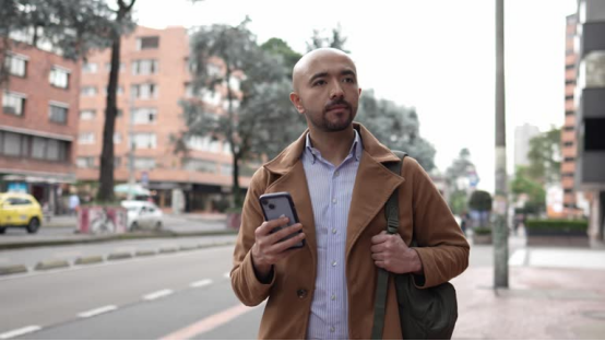 Bald man (with a smooth scalp maintained by a head shaver) standing on an urban street, holding a phone.