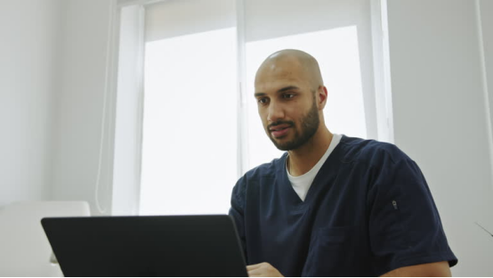 A professional in scrubs working at a laptop, representing the busy lifestyle that benefits from the convenience of a head shaver.